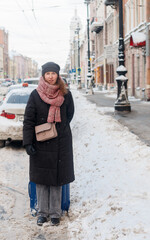 A pretty young woman in a hat and scarf stands with a blue suitcase  on a snowy road in a winter city waiting for a taxi. Selective focus