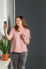 A pretty young woman with red hair in a light T-shirt and gray jeans stands against a gray wall, smiling and posing with a mobile phone. A flower on the window. Selective focus, copy space