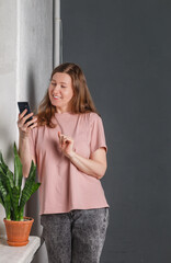 A pretty young woman with red hair in a light T-shirt and gray jeans stands against a gray wall, smiling and posing with a mobile phone. A flower on the window. Selective focus, copy space