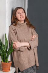 A pretty young woman with red hair in a beige sweater is smiling happily against a gray wall. A flower on the window. Selective focus