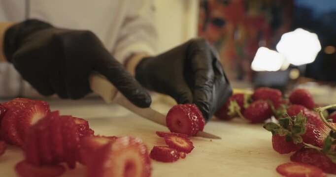 A professional chef wearing black nitrile gloves carefully slices ripe red strawberries with a sharp knife on a wooden cutting board in a warm, lit kitchen environment. High-quality culinary footage.
