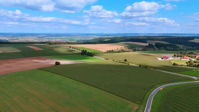 The scene shows a wide view of farmland with green fields and patches of brown soil. A winding road cuts through the landscape.