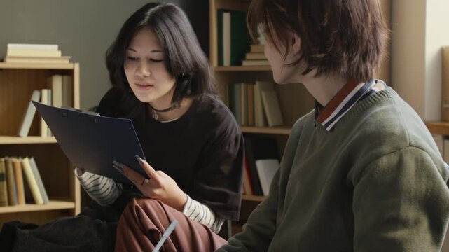 Tilt up shot of two teenage girls discussing mental health questionnaire reading questions on clipboard while participating in group therapy session in college counselling room