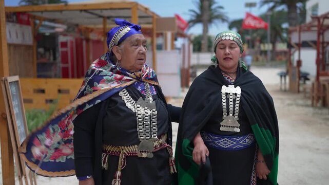 Two mapuche women wearing traditional clothes and silver jewelry walking along a sandy street
