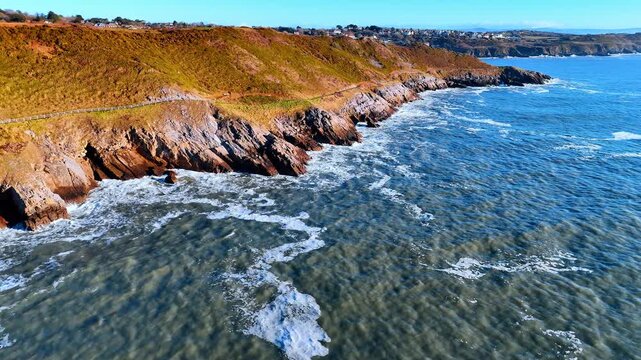 The view shows a coastline with rocks and waves crashing against them. The land rises gently from the shore, featuring greenery and a path along the edge.