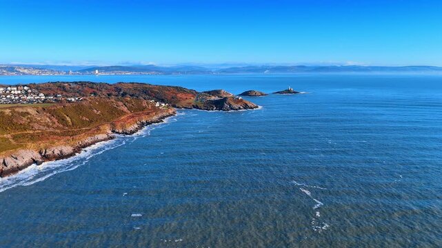 The scene shows a coastline with sandy beaches and waves rolling through the water. Land rises steeply along the shore, with buildings visible in the distance.