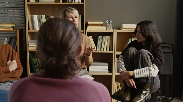 Panning shot of adult female psychotherapist talking to group of teenage students discussing emotions together during therapy session at college counselling office
