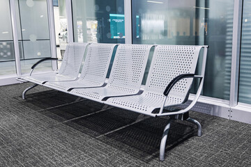 Empty metal waiting chairs in airport terminal. Modern perforated bench seats by glass wall. Public transport waiting area interior. Clean minimalist travel space. Nobody waiting.