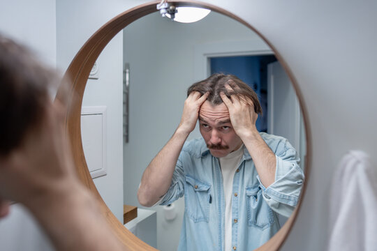 Worried young man examines thinning hair in bathroom mirror during morning routine. Self awareness, concern about appearance. Thoughtful male checks hairline. Men hair loss problem, male baldness.
