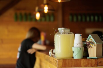 Bright lemonade fills a glass jar on a warm wooden counter, while a person prepares drinks in the...