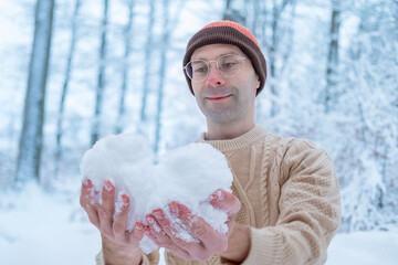 Romantic young man in beanie, sweater holds snow heart in winter forest, love, Valentine&rsquo;s Day, snowball in snowy forest, smiling camera, beauty heartfelt gestures in nature, active outdoor lifestyle