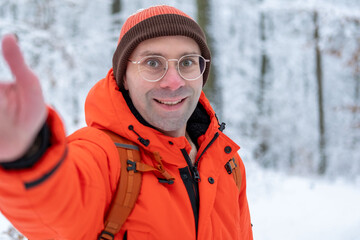Happy young Smiling man in bright orange jacket taking selfie in snowy forest, enjoying winter walk, outdoor lifestyle and freedom, Cheerful traveler taking selfie during winter walk in snowy woods