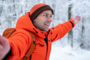 Happy young Smiling man in bright orange jacket taking selfie in snowy forest, enjoying winter walk, outdoor lifestyle and freedom, Cheerful traveler taking selfie during winter walk in snowy woods