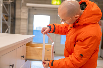 man in orange jacket measuring furniture with tape retail store, male customer ensuring correct size paper tape, self service furniture store warehouse aisle, home renovation, consumer shopping task