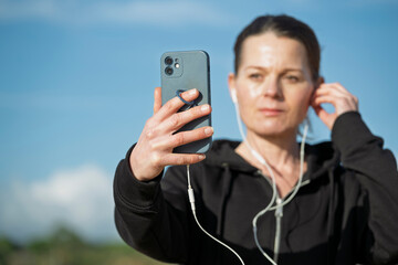 Sporty woman in a black hoodie taking a selfie with a smartphone while wearing white earbuds.