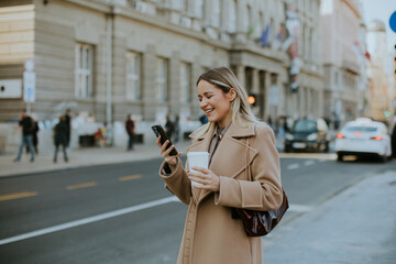 Fototapeta premium Woman holds coffee and checks phone on city street in autumn