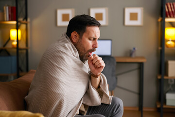 Unwell man feeling sick and coughing while sitting on a sofa with a blanket
