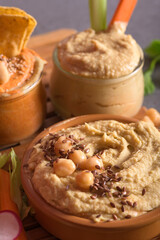 Assorted hummus bowls with chickpeas seeds and fresh vegetables on wooden boards over dark background
