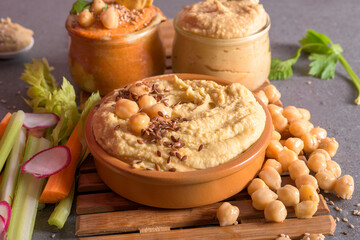 Assorted hummus bowls with chickpeas seeds and fresh vegetables on wooden boards over dark background