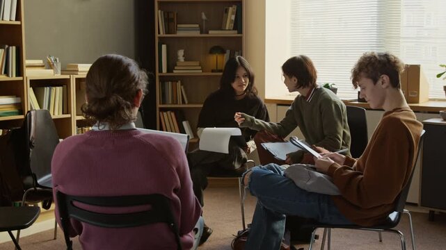 Group of young teenagers discussing mental health questionnaire completing test forms on clipboards while sitting on chairs in counselling room filled with sunlight