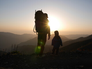 A father with backpack carrier and his child are hiking in mountains and looking at sunset. Not AI