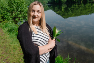 Young woman holding beautiful bouquet of a lillies of a valley flowers standing against forest lake landscape.
