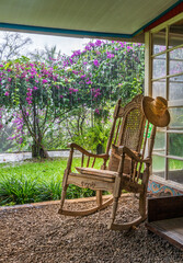 A vintage wooden rocking chair with a straw hat rests on a porch in a lush garden setting in Costa Rica. The scene evokes relaxation and vacation