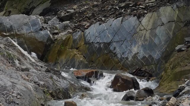Footage captures a small, fast-flowing mountain stream with white water cascading over dark, jagged rocks. The surrounding rock face features striking, geometric patterns, possibly columnar basalt, wi