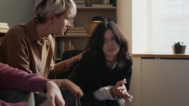 Distressed teenage girl with black hair covering face with hands sharing emotions with female therapist during shared psychotherapy session in counselling room