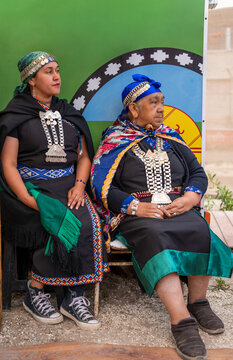 Mapuche women sitting with traditional clothing, textiles, and silver jewelry against a cultural flag