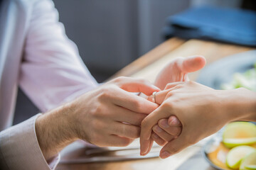 Man putting engagement ring on smiling woman's finger during romantic proposal at home with coffee