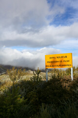 Translation: Military zone, no entry, danger of explosions. Information sign in a mountainous area, yellow in color, warning of the boundaries of a military exercise zone.