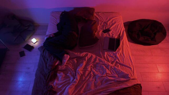 Man lying on messy bed at night using smartphone near laptop under colorful neon light, modern loneliness and digital lifestyle at home, insomnia, overthinking and online escape