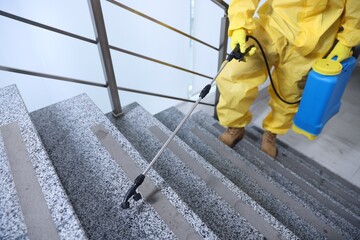 Pest control worker in protective suit spraying insecticide on stairs indoors, selective focus. Space for text