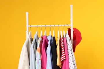 Different clothes and baseball cap hanging on rack against orange background, closeup