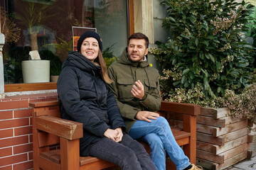 Man and woman sitting together on wooden bench near building and plants