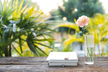 Stack of notebook and vase of rose on wooden table with backyard nature view