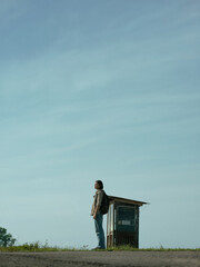 Person standing near a small rural bus stop under a wide open sky. Minimalist composition, quiet countryside atmosphere, everyday travel and waiting concept. AI generated illustration.