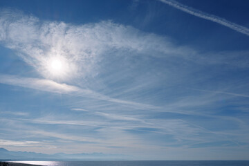 Clear blue sky over calm sea with bright sun and wispy clouds, expansive horizon and reflective...