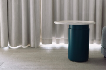 Round table on a blue pedestal base stands in a minimal living room with light curtains and soft...