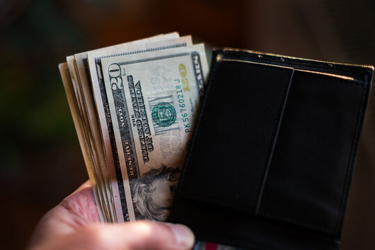 A blurred hand places or taking folded US dollar bills, with 20$ banknote visible, into a dark wallet. Concepts of savings, payment, budget, and finance