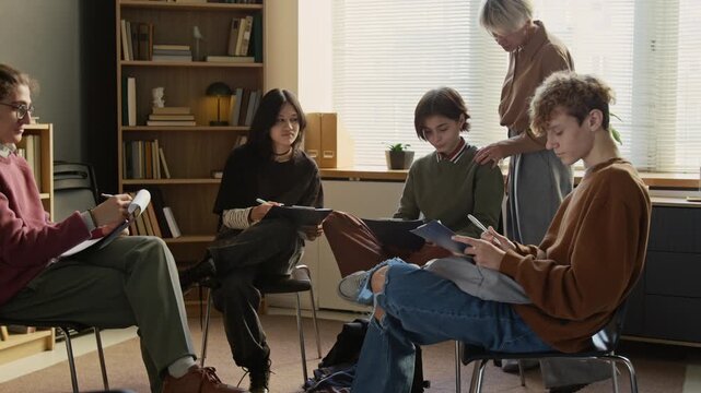 Middle aged female therapist supervising group of teenagers completing mental health questionnaires on clipboards during session in campus counselling room
