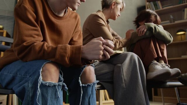 Cropped shot of anxious teenage boy fidgeting with fingers clasping palms while female psychotherapist comforting teenage girl during group therapy session