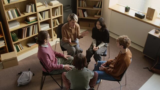 High angle shot of middle aged female psychotherapist sitting among circle of teenagers holding hands offering mutual support at end of group therapy session in counselling room