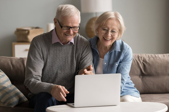 Studying digital world at golden age. Happy senior couple pensioners hug on sofa using notebook at improvised home office enjoy watching webinar tutorial online take hobby course on elearning platform