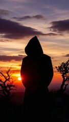 The figure of a hooded monk stands out in silhouette against the sunset landscape.