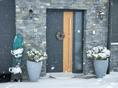Modern alpine house entrance framed by natural stone walls during heavy snowfall. Planters with snowy pine trees, snowboard and wooden door with wreath create cozy winter atmosphere in the countryside