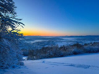 Naklejka premium Winter panorama of a snowy valley and distant hills during blue hour at sunrise. Soft light spreads across forests and countryside, blending deep blues with warm tones for a tranquil winter scenery.