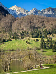 Fototapeta premium Tranquil alpine landscape with a green spring meadow and calm lake below rugged mountain peaks covered with snow. Seasonal transition from winter to spring in a peaceful mountain valley in the Alps.