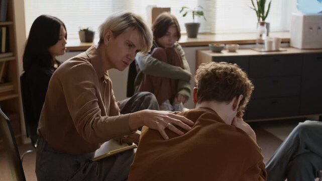 Adult female psychotherapist comforting distressed teenage boy guiding and supporting young participants in therapeutic group session in counselling room